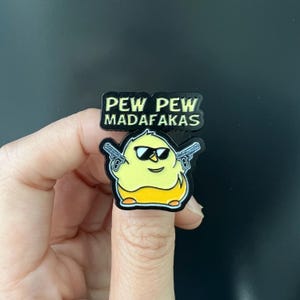 Puede incluir: Un pin con forma de pollito amarillo con gafas de sol y dos pistolas. El pin tiene el texto "PEW PEW MADAFKAS" en letras negras. El pollito tiene un cuerpo amarillo y pies naranjas.