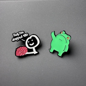 May include: Two enamel pins on a grey surface. One pin features a stick figure with a brain and the text "Hey, You dropped this". The other pin is a green frog with its arm raised.