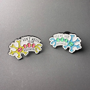May include: Two enamel pins with neuron designs. One pin is yellow with the text "DON'T WASTE YOUR POTENTIAL". The other is blue with the text "YOU'VE GOT POTENTIAL". Both pins have a white outline and black backing.
