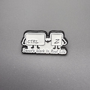 May include: A black and white enamel pin featuring two rectangular keyboard keys labeled CTRL and Z with stick-figure arms and legs. The text below the keys reads, "Doesn't Work In Real Life," written in a cursive font on a white background.