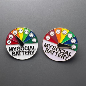 Puede incluir: Dos pines esmaltados con diseño de indicador. El indicador está dividido en segmentos de colores del arcoíris, cada uno con una cara de emoji diferente. El texto "MY SOCIAL BATTERY" está impreso en la parte inferior de cada pin.