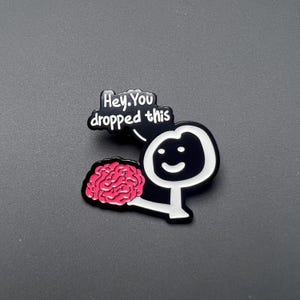 Peut inclure: Épingle en émail noir et blanc représentant un bonhomme allumette tenant un cerveau rose. L'épingle comprend une bulle de dialogue avec le texte "Hey, You dropped this". Le design est sur fond gris.