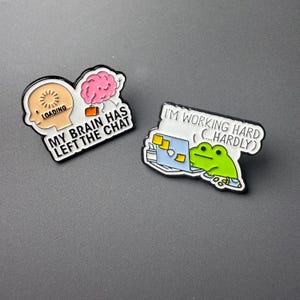 Könnte beinhalten: Zwei Emaille-Anstecknadeln mit schwarzen Rändern. Eine Anstecknadel zeigt einen Kopf mit einem Ladesymbol und dem Text "MY BRAIN HAS LEFT THE CHAT". Die andere zeigt einen Frosch, der an einem Laptop arbeitet, mit dem Text "I'M WORKING HARD (...HARDLY)."