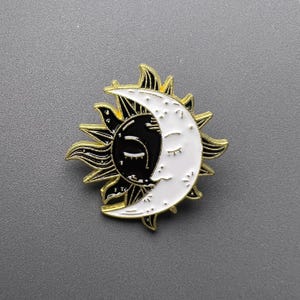 Op de afbeelding: Een emaille pin met een hemels thema, met een zon en een maan. De pin heeft een goudkleurige omtrek, met de zon half zwart en de maan half wit. De zon en de maan kijken elkaar aan, met het gezicht van de maan gesloten in slaap.