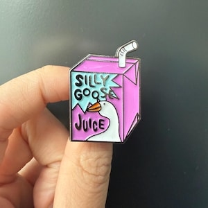 May include: A pink enamel pin shaped like a juice box with a white goose and the words "Silly Goose Juice". The juice box has a blue background and a white straw. The pin is a fun accessory.