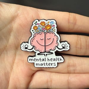 Puede incluir: Un pin de esmalte rosa con forma de cerebro sonriente con flores y la frase "mental health matters". El diseño es blanco y negro. El cerebro sonríe y tiene flores.