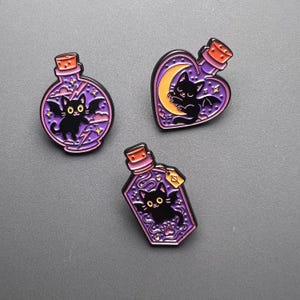 May include: Three enamel pins featuring black cats inside purple potion bottles. One pin is round with a bat-winged cat, another is heart-shaped with a cat on a crescent moon, and the third is a pentagon shape.