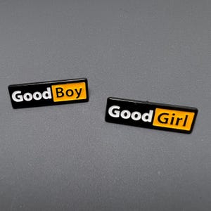 May include: Two rectangular enamel pins. One pin reads "Good Boy" in white text on a black background with a yellow rectangle. The other pin reads "Good Girl" in white text on a black background with a yellow rectangle.