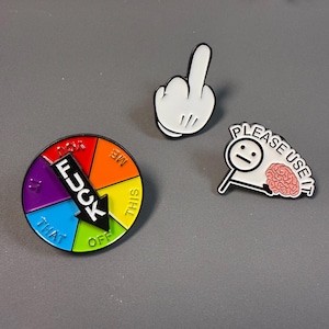 May include: Three enamel pins on a gray surface. One pin is a white hand giving the middle finger. Another is a colorful wheel with the word "FUCK" and an arrow. The third pin has a brain and the words "PLEASE USE IT".