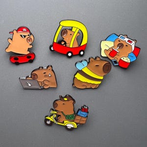 Peut inclure: Collection d'épingles en émail sur le thème du capybara. Un capybara fait du skateboard, un autre conduit une voiture rouge et jaune, et un troisième porte des lunettes 3D. D'autres épingles incluent un capybara en abeille, travaillant sur un ordinateur portable et à scooter.