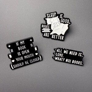 May include: Three black and white enamel pins with book-themed text. One pin shows a character in bed reading with the text SLEEP IS GOOD... BOOKS ARE BETTER. Another reads IF MY BOOK IS OPEN YOUR MOUTH SHOULD BE CLOSED. The third reads ALL WE NEED IS.. LOVE. WHAT? NO! BOOKS.