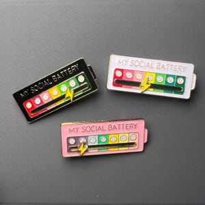 May include: Three enamel pins with the text "MY SOCIAL BATTERY". Each pin features a battery-style design with a range of emoji faces and a lightning bolt. The pins are in black, white, and pink with varying color schemes.