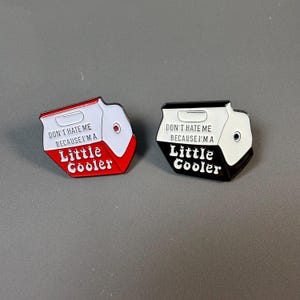 May include: Two enamel pins shaped like coolers. One is red, white, and black, and the other is black and white. Each pin has the text "DON'T HATE ME BECAUSE I'M A Little Cooler" in white lettering.