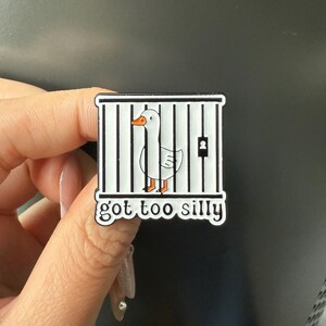 May include: A square enamel pin featuring a white goose inside a black and white cage. The pin has the text "got too silly" in a black font. The goose has an orange beak and feet.