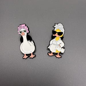 Puede incluir: Dos pines de dibujos animados con un ganso y un pato. El ganso lleva rulos rosas y gafas de sol. El pato lleva una bata y una toalla blancas, también con gafas de sol. Ambos tienen detalles en negro y pies rosas.