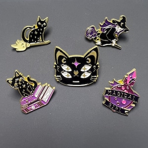 Op de afbeelding: Een verzameling van vijf emaille pins met zwarte katten en heksen thema's. Ontwerpen omvatten een kat op een bezem, een kat met meerdere ogen, een kat met boeken en een 'Magical Girl' drankje.