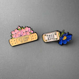 Puede incluir: Dos pines de esmalte. Uno es una tirita con flores rosas y el texto "IT'S GONNA BE OKAY." El otro es un frasco de pastillas con una flor azul y el texto "STRUGGLING DOES NOT MEAN YOU ARE FAILING."