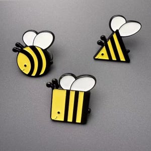 May include: Three enamel pins shaped like bees, each featuring a distinct geometric body: one round, one triangular, and one square. Each pin displays yellow and black stripes, white wings, and a small black dot for an eye on a gray background.