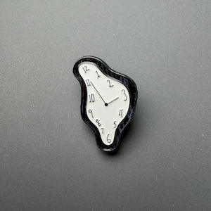 May include: A black and white enamel pin shaped like a melting clock with an irregular, fluid outline. The white face features black numerals from one to twelve arranged in a distorted circle, with two black hands pointing to the time.