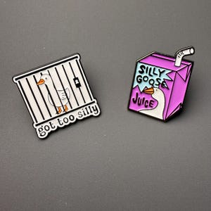 May include: Two enamel pins featuring a cartoon goose. One pin shows a goose in a cage with the text "got too silly". The other pin is a juice box with a goose and the text "Silly Goose Juice".
