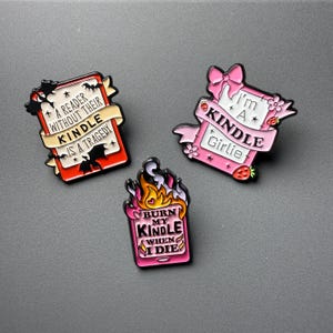 May include: Three enamel pins with text and graphics. One pin is red with the text "A READER WITHOUT THEIR KINDLE IS A TRAGEDY". Another is pink with "I'm A Kindle Girlie". The third is pink with flames and "BURN MY KINDLE WHEN I DIE".