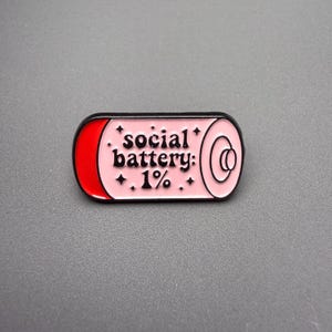 Op de afbeelding: Een rechthoekige emaille speld in de vorm van een batterij met afgeronde randen. De speld heeft een lichtroze middenstuk met zwarte tekst die social battery: 1% leest, omringd door kleine zwarte sterretjes. Links bevindt zich een rood vlak.