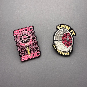 May include: Two enamel pins. The left pin is rectangular with the word STATIC repeated in pink, featuring a pink car wheel and suspension strut. The right pin is circular with the text SPIN IT BURN IT, featuring a white and red car wheel design.