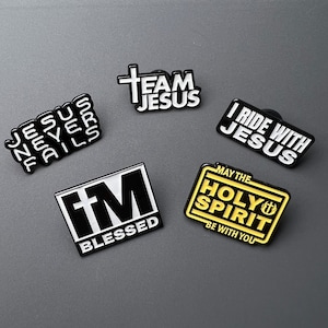 May include: A collection of black and white enamel pins with religious phrases. The pins include text such as "Jesus Never Fails", "Team Jesus", "I Ride With Jesus", "Blessed", and "May The Holy Spirit Be With You".