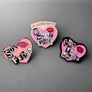 May include: Three enamel pins with pink heart shapes and black borders. One pin says "So Fetch", another says "On Wednesdays We Wear Pink", and the third says "Boo You Whore". Each pin features a red lip design.