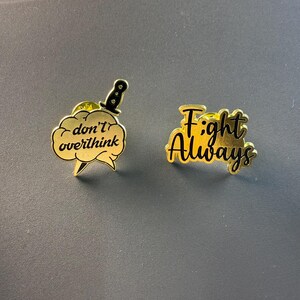 Könnte beinhalten: Zwei goldfarbene Emaille-Anstecknadeln. Eine Nadel zeigt ein Messer durch eine Wolke mit den Worten "don't overthink". Die andere Nadel sagt "Fight Always" in einer stilisierten Schrift. Beide Nadeln haben schwarze Akzente und liegen auf einer grauen Oberfläche.