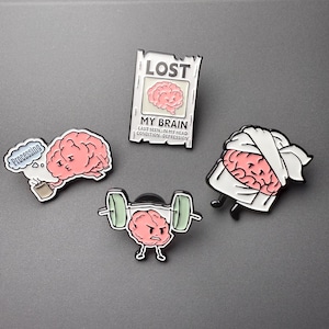 May include: Four enamel pins featuring pink brain characters. One is a lost poster reading LOST MY BRAIN. Another shows a brain holding a coffee mug with a speech bubble saying Processing. A third brain lifts a barbell. The fourth wears a white towel.
