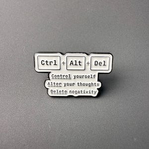 Puede incluir: Pin de esmalte blanco y negro con forma de teclas de ordenador. El pin presenta el texto "Ctrl + Alt + Del" sobre las palabras "Control yourself, Alter your thoughts, Delete negativity."