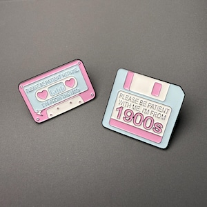 May include: Two enamel pins featuring a light blue and pink colour scheme. One pin is shaped like a cassette tape with the text PLEASE BE PATIENT WITH ME. I'M FROM THE 1900s. The other pin is a floppy disk with the text PLEASE BE PATIENT WITH ME. I'M FROM 1900s.