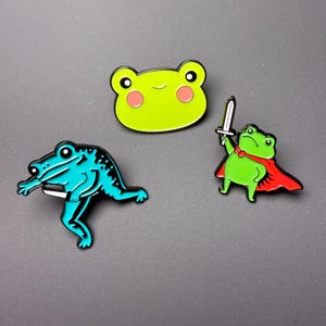 May include: Three enamel pins featuring frog designs. One is a lime green frog face with pink cheeks. Another is a bright blue frog in a dynamic pose holding a small knife. The third is a green frog wearing a red cape and holding a silver sword.