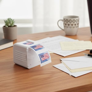 May include: A white stamp dispenser with American flag stamps on a wooden desk. A laptop, pen, mug, and envelopes are also on the desk. A small succulent plant in a pot is in the background.