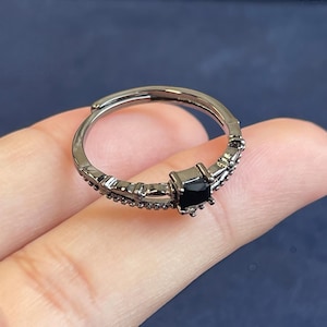 Puede incluir: Anillo negro ajustable con una piedra preciosa negra cuadrada en el centro. El anillo tiene un acabado metálico oscuro y está adornado con pequeños detalles decorativos a lo largo de la banda. Fondo azul oscuro.
