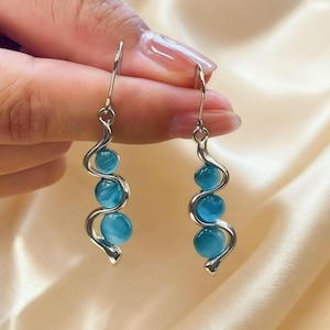 May include: A pair of dangle earrings with a silver-tone, wavy design. Each earring features three round, light blue beads. The earrings are suspended from a simple hook.