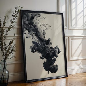 Puede incluir: Una impresión artística enmarcada con una nube de tinta negra sobre un fondo crema. El marco negro se apoya en una pared blanca y un suelo de madera. Un jarrón de cristal con ramas secas está a la izquierda.