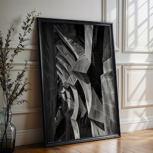 May include: A black and white framed print of an architectural design. The artwork shows an abstract composition of angular shapes and contrasting tones. The black frame leans against a white wall and wooden floor, creating a minimalist aesthetic.
