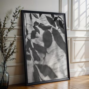 Könnte beinhalten: Ein gerahmter Schwarz-Weiß-Druck von Blätterschatten, der an einer weißen Wand lehnt. Das Kunstwerk befindet sich in einem schwarzen Rahmen und steht auf einem Holzboden. Links vom Kunstwerk befindet sich eine Glasvase mit Zweigen.
