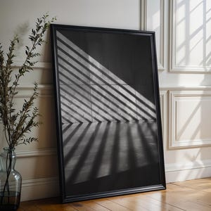 Puede incluir: Una gran obra de arte enmarcada en negro se apoya contra una pared, con luz y sombra creando un patrón geométrico en su superficie. Un jarrón de cristal con ramas está cerca. El marco oscuro de la obra contrasta con la pared clara.