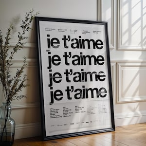 May include: A framed poster with the phrase "je t'aime" repeated in bold black letters on a white background. The poster is in a black frame and leans against a white wall. A vase with branches is to the left.