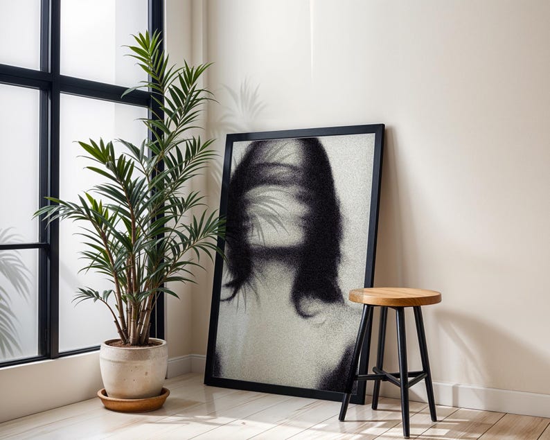 Puede incluir: Una impresi&oacute;n art&iacute;stica enmarcada en negro con un retrato en blanco y negro de una mujer con rasgos faciales borrosos. Un peque&ntilde;o taburete de madera con patas negras est&aacute; cerca. Una planta en maceta con hojas verdes est&aacute; frente a una ventana.