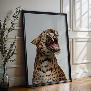 May include: A framed art print depicting a roaring leopard. The leopard's distinctive spotted coat is prominent, and its open mouth reveals sharp teeth. The artwork is framed in a black border and rests against a white wall.