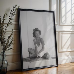 Könnte beinhalten: Schwarz-weiß gerahmte Fotografie einer Frau in einem weißen Kleid, die sitzt und eine Schüssel hält. Das Foto lehnt an einer weißen Wand, neben einer Glasvase mit Zweigen.