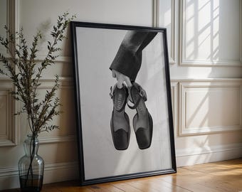 Puede incluir: Una impresión de arte de pared enmarcada en blanco y negro que muestra un par de zapatos de cuero negro con correa al tobillo y punta cuadrada. Los zapatos son sostenidos por una mano con manga oscura, y el texto JEU D'ART es visible en las plantillas.