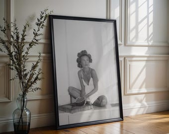 Op de afbeelding: Een zwart-wit ingelijste kunstprint met een persoon met haarkrullers die op een oppervlak zit, een lichtgekleurde jurk met spaghettibandjes draagt en een kleine kom vasthoudt. Het kunstwerk is gevat in een eenvoudige zwarte lijst met dunne rand.