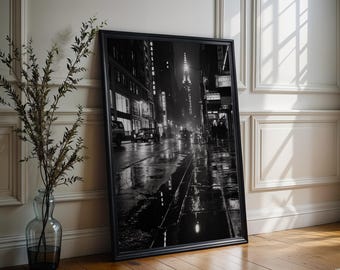 May include: A black and white framed wall art print depicting a rainy New York City street scene at night. The image features wet pavement with reflections, city buildings, illuminated signs, and a prominent skyscraper in the distance.