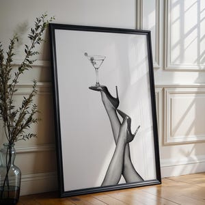 Może przedstawiać: Czarno-biały oprawiony wydruk artystyczny przedstawiający kieliszek do martini zrównoważony na wysokich obcasach. Grafika przedstawia parę nóg w szpilkach trzymających martini z oliwkami. Wydruk jest prezentowany na drewnianej podłodze.