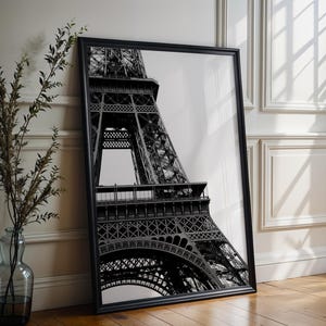Puede incluir: Impresión enmarcada en blanco y negro de la Torre Eiffel. La fotografía captura una vista detallada de la intrincada estructura de celosía de hierro de la torre. El arte enmarcado se apoya en una pared blanca y un suelo de madera.